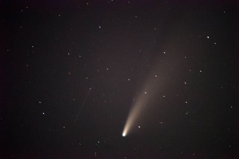 Comet NEOWISE, a green-white head trailing a long pale dust tail across a dark starry sky, a faint streak at left