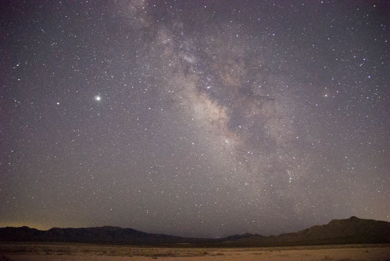 Milky Way core rising over dark desert mountains, one bright point of light at left, faint orange glow on the horizon