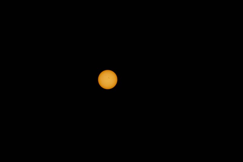 Orange disc of the sun through a solar filter, a tiny dark sunspot near its center, isolated against pure black