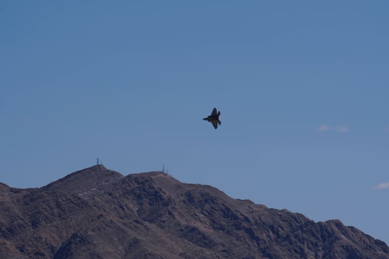 F-22 Raptor banking with afterburner glow high above a desert ridge dotted with antenna towers, deep blue sky