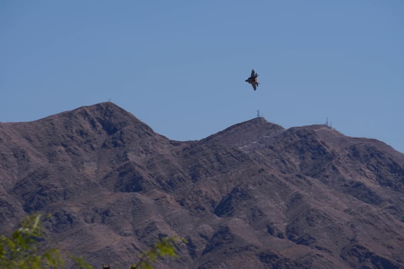 F-22 Raptor in a tight afterburner turn low over brown desert mountains topped with antenna towers