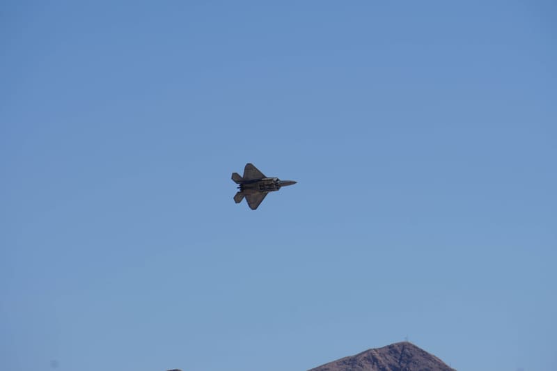 F-22 Raptor banking to show its underside with weapons bay doors open against a clear blue sky, a mountain peak below