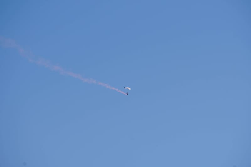 Skydiver under a white parachute canopy descending through blue sky, a pink smoke trail streaming behind