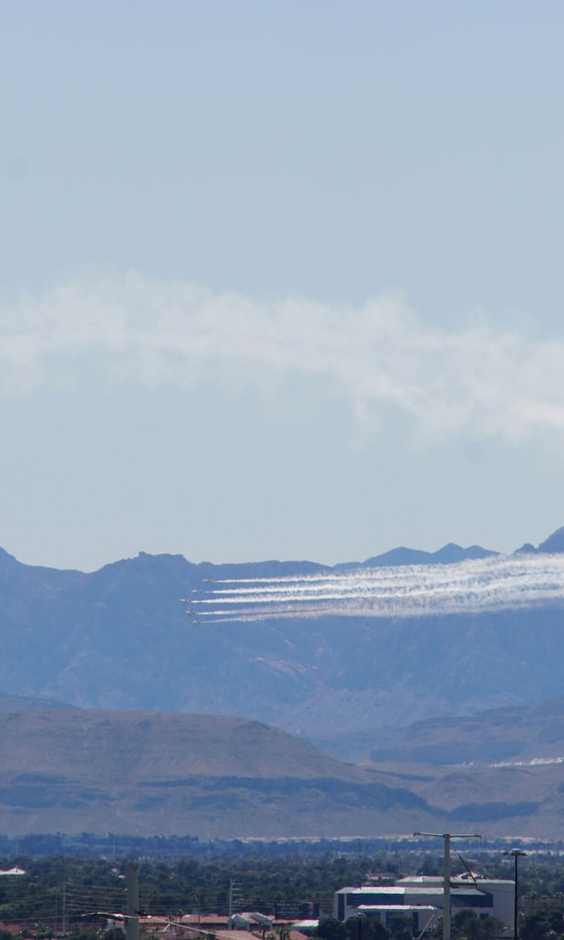 Six USAF Thunderbirds F-16s in delta formation trail white smoke low along hazy desert mountains above suburban rooftops
