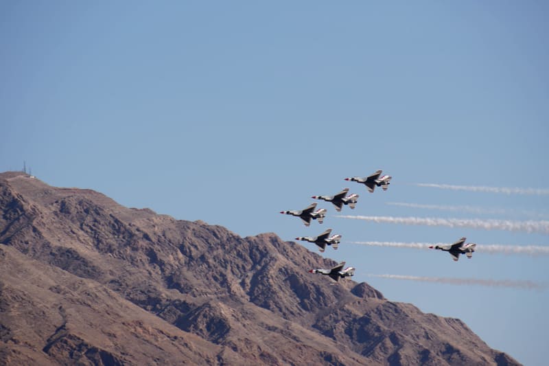 Six USAF Thunderbirds F-16s in delta formation skimming a brown desert mountain ridge, white smoke trails behind