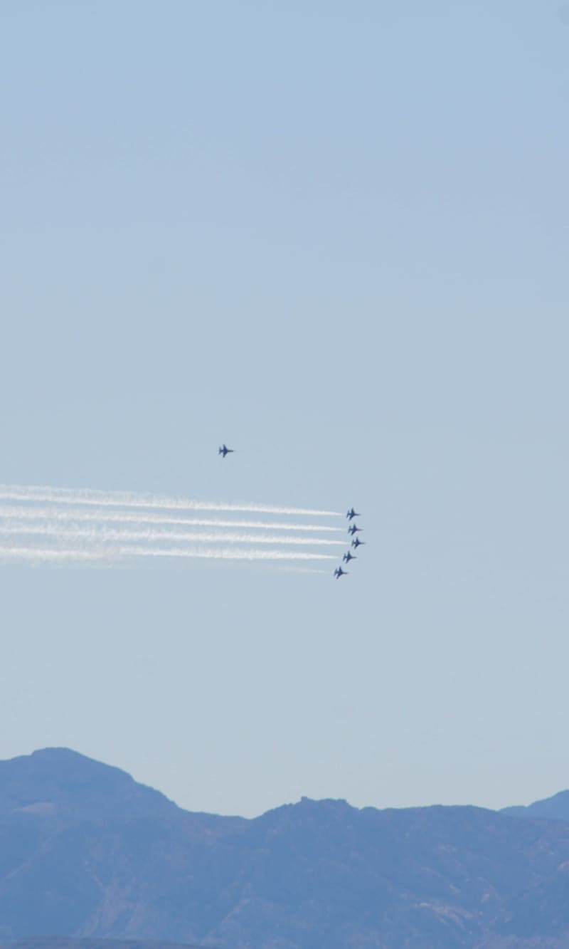 Five USAF Thunderbirds F-16s stacked in a column trailing white smoke, a solo jet above, over a dark mountain ridge