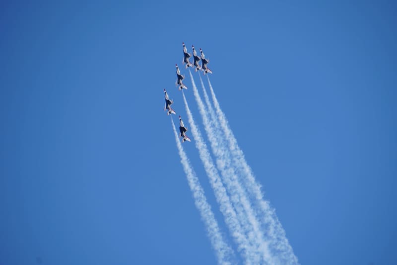 Six USAF Thunderbirds F-16s in a tight delta formation climbing steeply into a clear blue sky, trailing white smoke