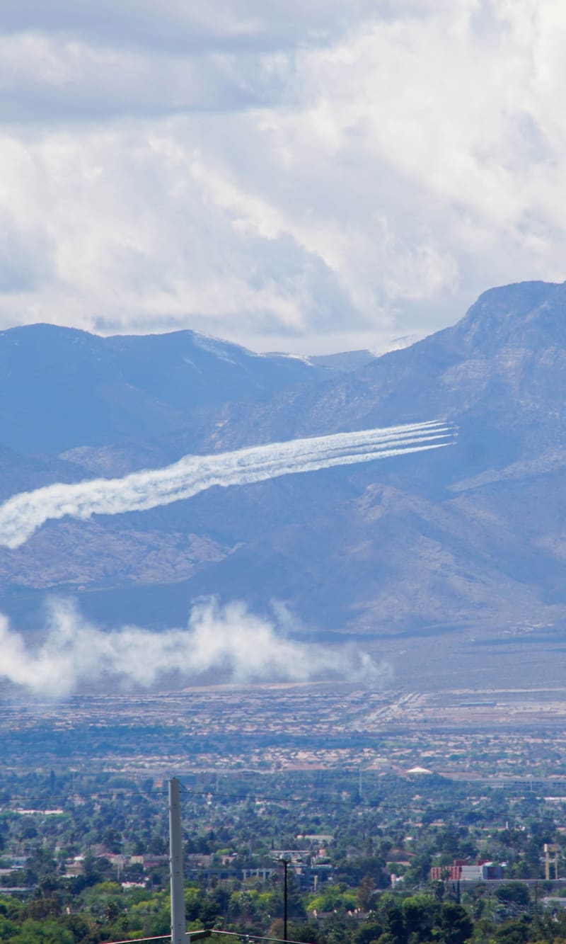 Four USAF Thunderbirds F-16s streak white smoke trails across cloud-capped mountains above a suburban valley