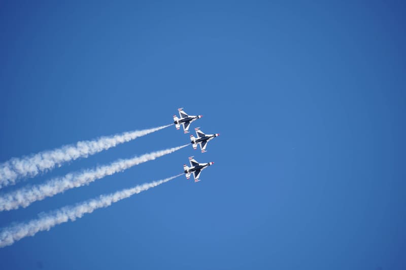 Three USAF Thunderbirds F-16s stacked in a tight diagonal line, white smoke trails streaming left across deep blue sky