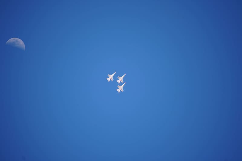 Three USAF Thunderbirds F-16s in a tight triangle, small against deep blue sky with a pale daytime moon in the upper left