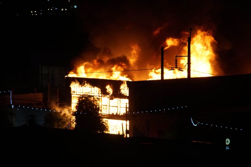 Two-story building burning at night, flames pouring from every window and over the roof, a tree silhouetted in front