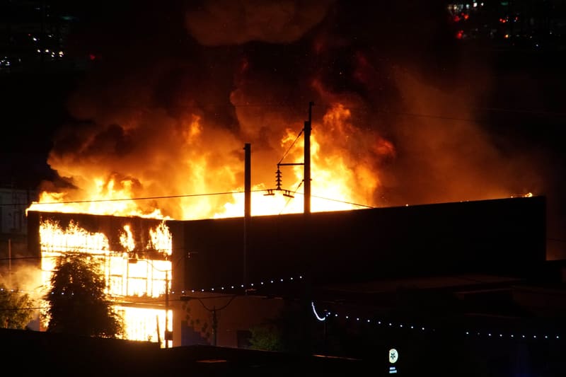 Huge orange flames and a thick smoke plume engulfing a two-story building at night, a utility pole silhouetted in front