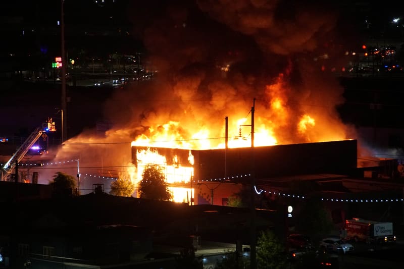 Fire truck aerial ladder raised beside a burning building at night, flames and a towering smoke plume over parked cars