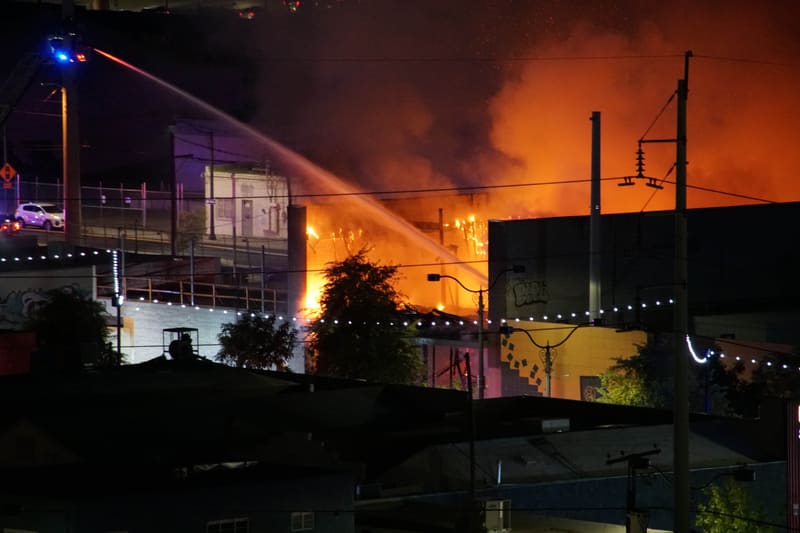 Fire hose stream arcing over a burning building at night, orange flames and smoke behind power lines and string lights