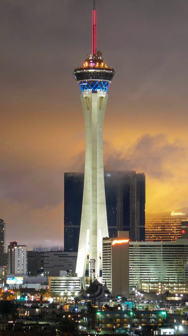 The Strat tower lit blue and red at night, its white legs rising past dark glass towers into an orange, hazy cloud bank