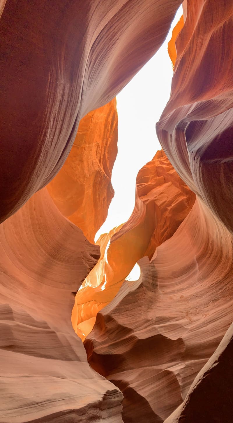 Fiery orange chamber glowing deep in Antelope Canyon, framed by curving salmon sandstone walls under a slit of white sky