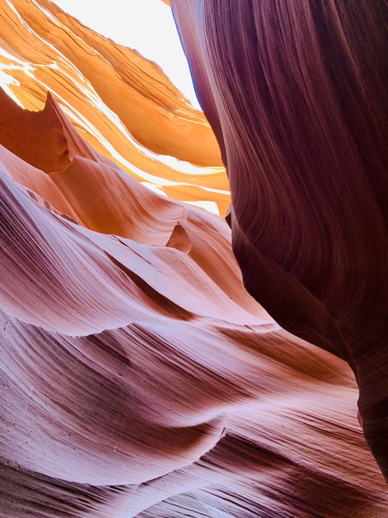 Wavy pink sandstone striations flow across Antelope Canyon below a glowing orange wall and a sliver of blue sky