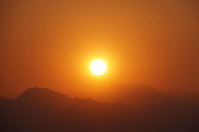 Bright white sun sinking into a thick orange haze above layered silhouettes of desert mountain ridges