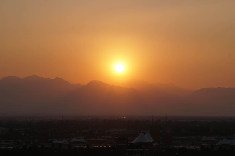 Hazy orange sunset over desert mountains, with city buildings, light poles and white tent-style roofs silhouetted below