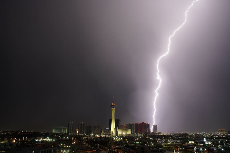 Thick blazing lightning bolt striking behind a red-lit tower beside the Strat, lighting Las Vegas storm clouds