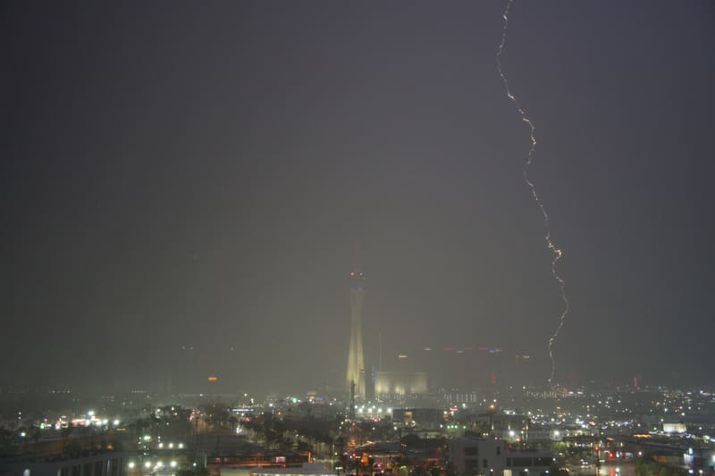 Thin orange-tinted lightning bolt drops through heavy storm haze to the right of a dimly lit Strat tower in Las Vegas