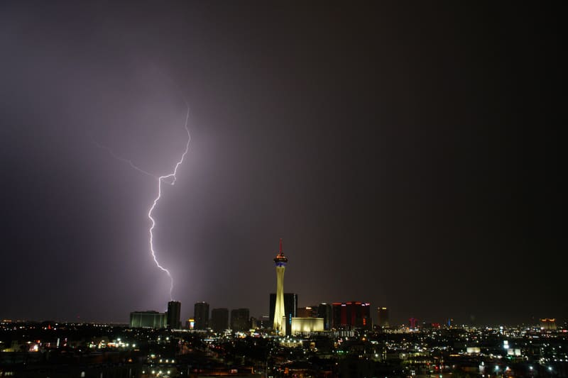 Forked lightning bolt striking down behind the skyline left of the glowing Strat tower under a purple Las Vegas sky