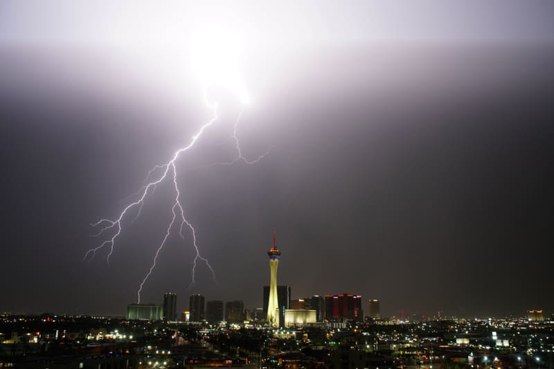 Lightning bursting from a glowing cloud base into many forked branches above the Las Vegas skyline, left of the Strat
