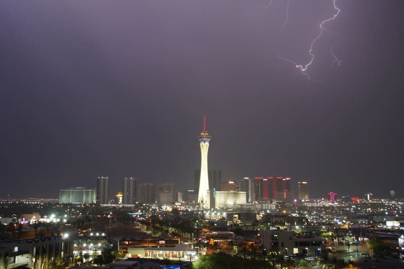 Forked lightning in the top right corner of a hazy purple-grey sky over the Strat tower and Las Vegas Strip hotels at night