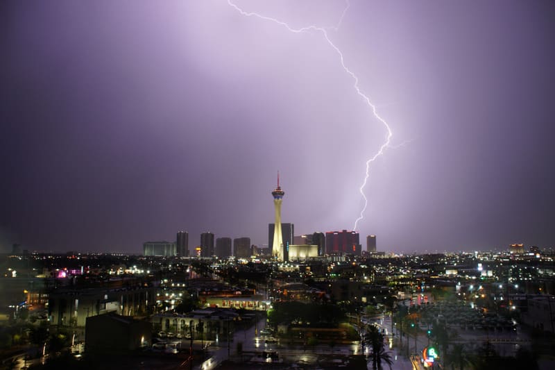Forked lightning bolt strikes down to the right of the Strat tower under a purple sky, wet Las Vegas streets glistening below