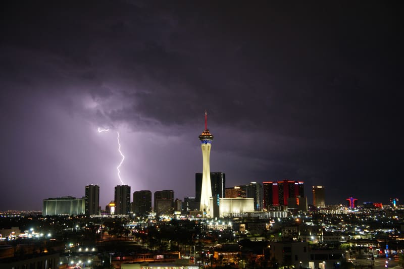 Jagged lightning bolt drops from purple storm clouds left of the Strat tower over the glowing Las Vegas skyline