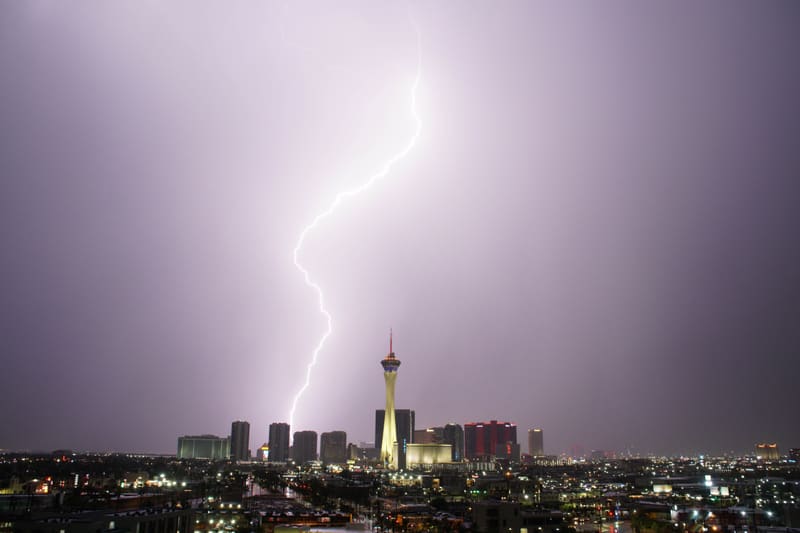 Bright jagged bolt striking the skyline left of the Strat tower under a glowing lavender sky over Las Vegas
