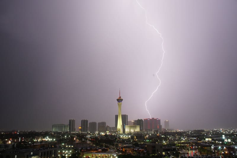 Bright lightning bolt drops straight down behind the red-lit towers beside the Strat, lavender storm sky over Las Vegas