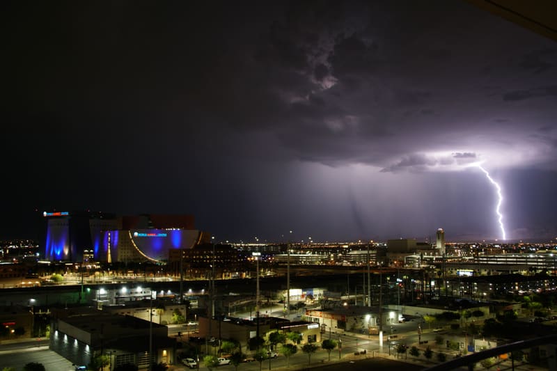 Lightning bolt strikes the horizon right of downtown Las Vegas at night, blue-lit World Market Center under purple clouds