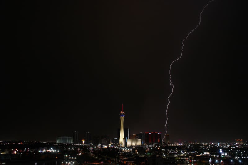 Slender lightning bolt cutting diagonally down a black sky to strike behind a red-lit tower right of the Strat in Las Vegas