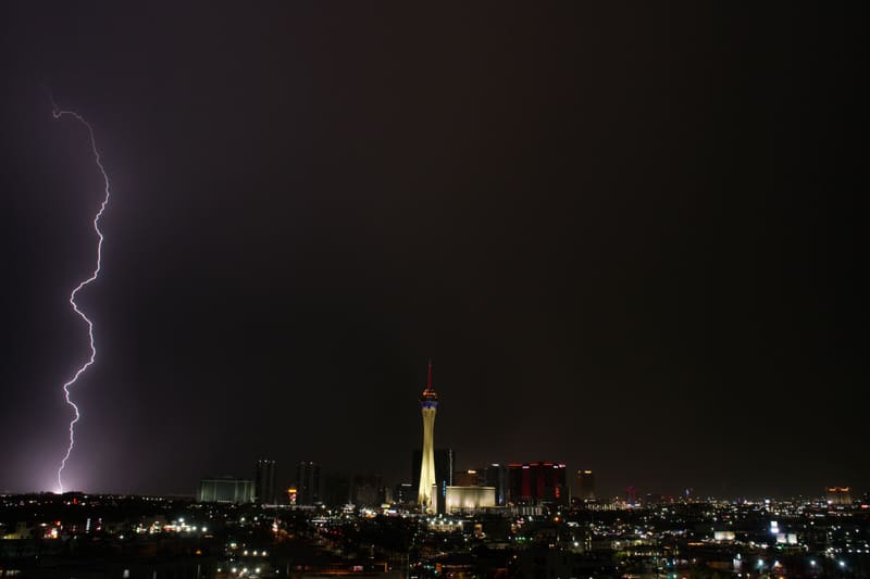 Thin zigzag lightning bolt at the far left edge of a black sky, the Strat tower glowing gold over the Las Vegas skyline