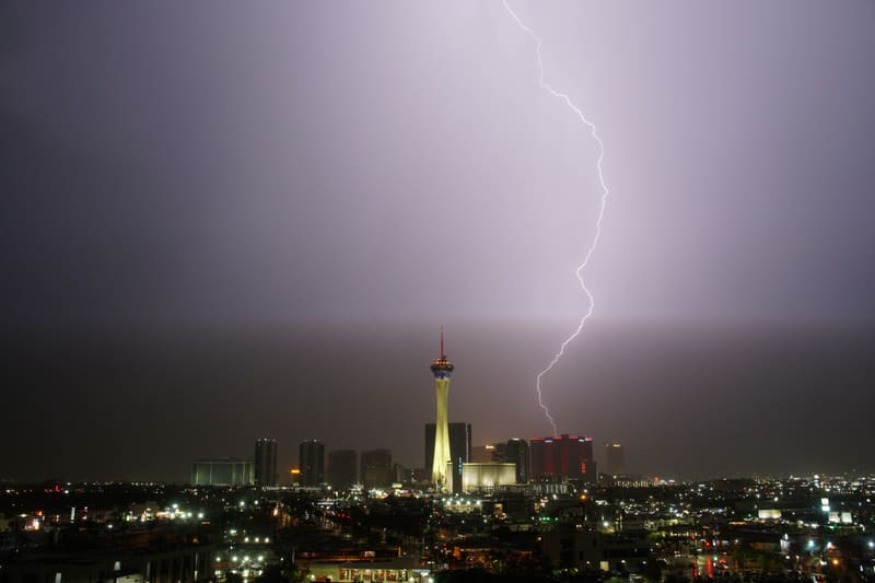 Single vertical lightning bolt strikes behind red-lit towers to the right of the Strat, Las Vegas skyline at night