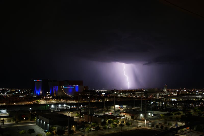 Thin vertical lightning bolt drops from a dark cloud base to the Las Vegas horizon beside a rain shaft, city lights below