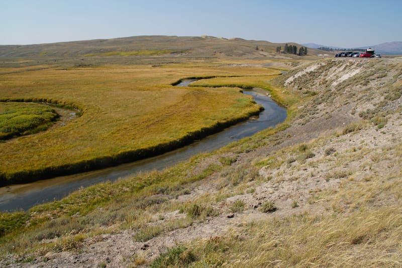 Slow river curving through flat golden grassland under a hazy blue sky, dry hillside in front and cars at a far pullout